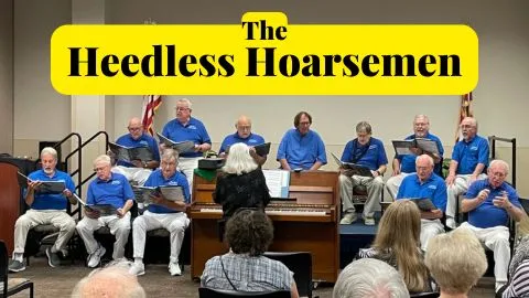 A photo of dozen men standing on a stage wearing matching sky-blue polos with header text that reads Heedless Hoarsemen