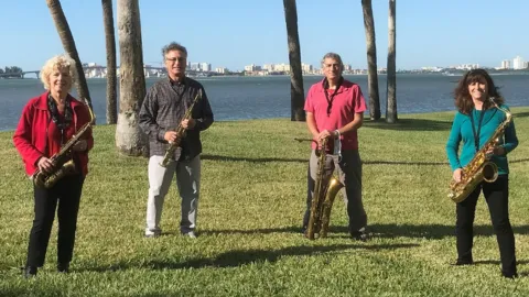 Four musicians stand outdoors on a grassy lawn near a waterfront, each holding a different type of saxophone. Palm trees surround them, and a cityscape is visible across the water under a clear blue sky.