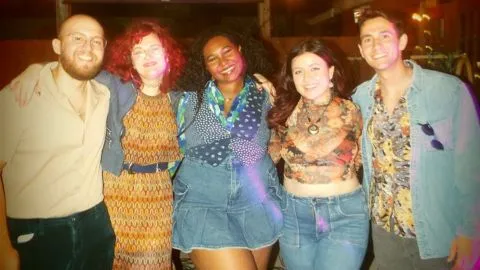 Five people stand close together outdoors at night, smiling warmly for the camera. From left to right: a bald man with a beard in a navy shirt and jeans; a woman with curly red hair in a patterned dress; a woman with voluminous dark curls wearing a colorful scarf and denim outfit; a woman with wavy dark hair in a floral top and jeans; and a man with short brown hair in a floral shirt and denim jacket. The group appears cheerful and relaxed, suggesting a casual or celebratory gathering.
