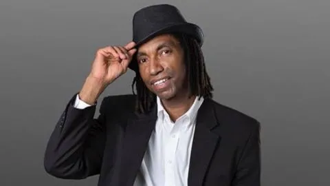 A man wearing a black blazer, white shirt, and black fedora tilts his hat and smiles warmly at the camera against a plain gray background.
